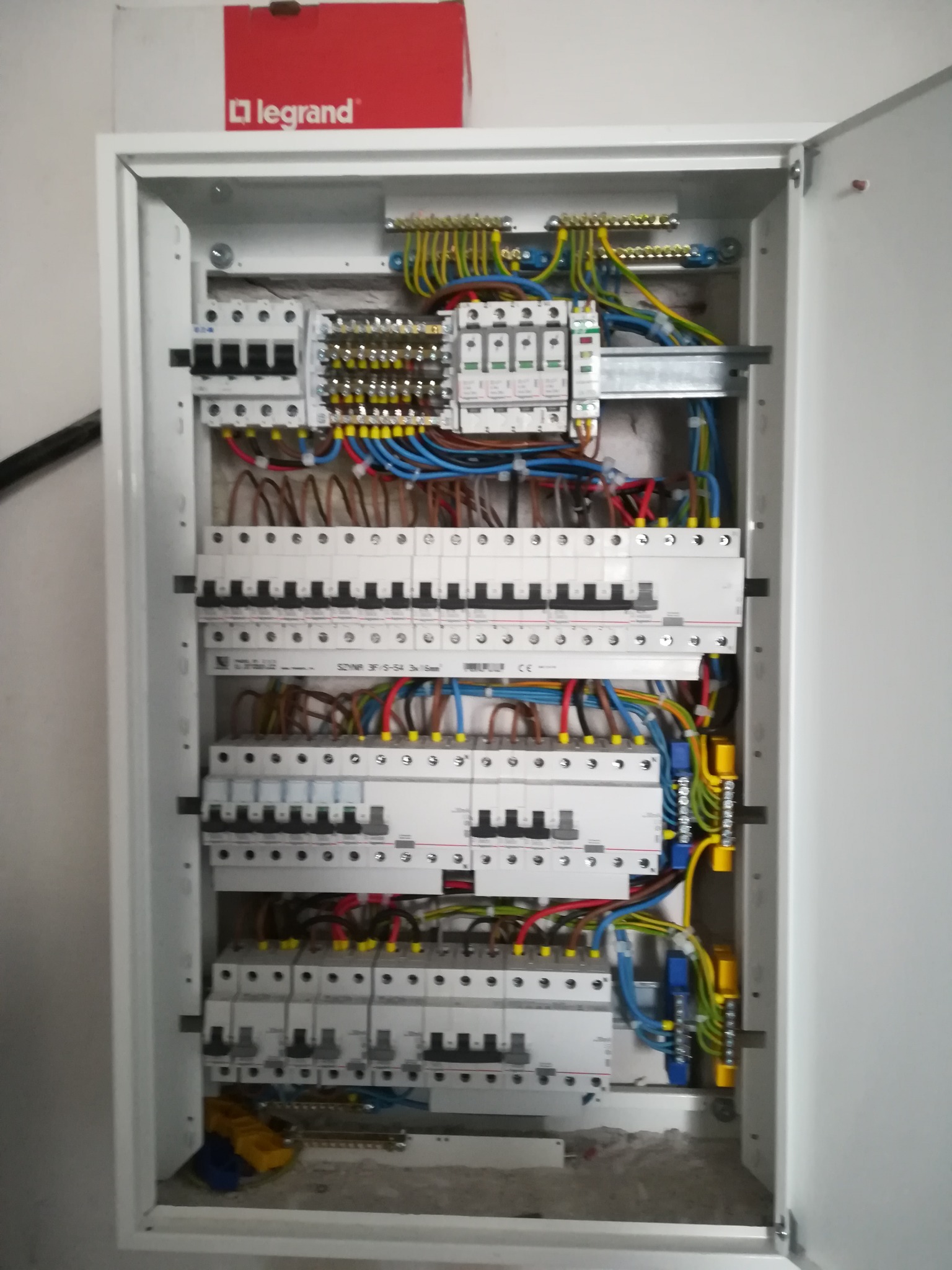 Otwarta biała szafa rozdzielcza z zainstalowanymi bezpiecznikami, przewodami i elementami łączeniowymi; widoczna czerwona skrzynka Legrand na górze.