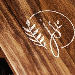 GRAFKA STUDIO IZABELA KARP - Drewniana powierzchnia z wygrawerowanym białym logo w kształcie inicjałów z motywem roślinnym, oświetlona ciepłym światłem.