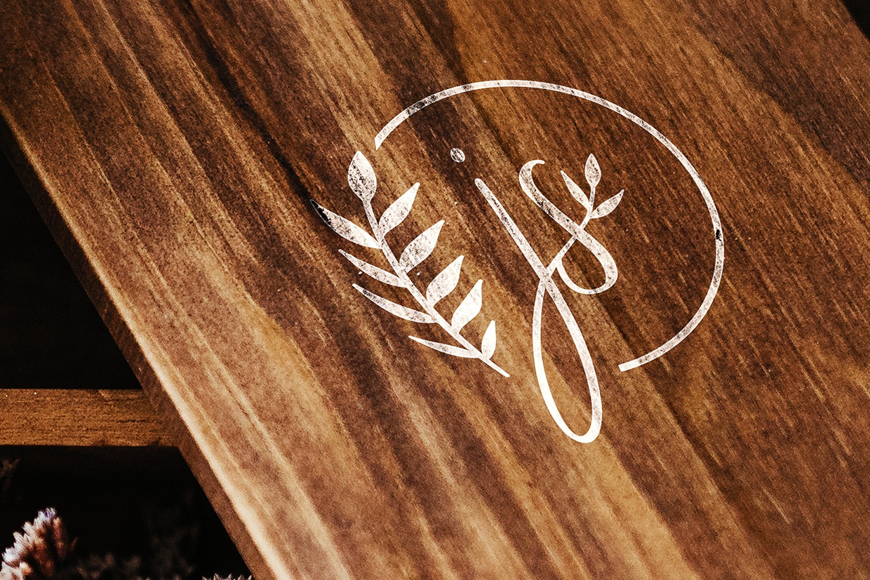Drewniana powierzchnia z wygrawerowanym białym logo w kształcie inicjałów z motywem roślinnym, oświetlona ciepłym światłem.