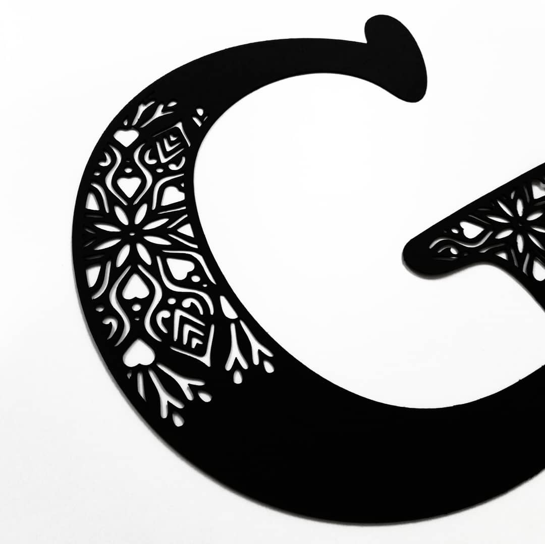 Dekoracyjna, ażurowa litera G z czarnego metalu, zdobiona motywami kwiatowymi i geometrycznymi, na białym tle.