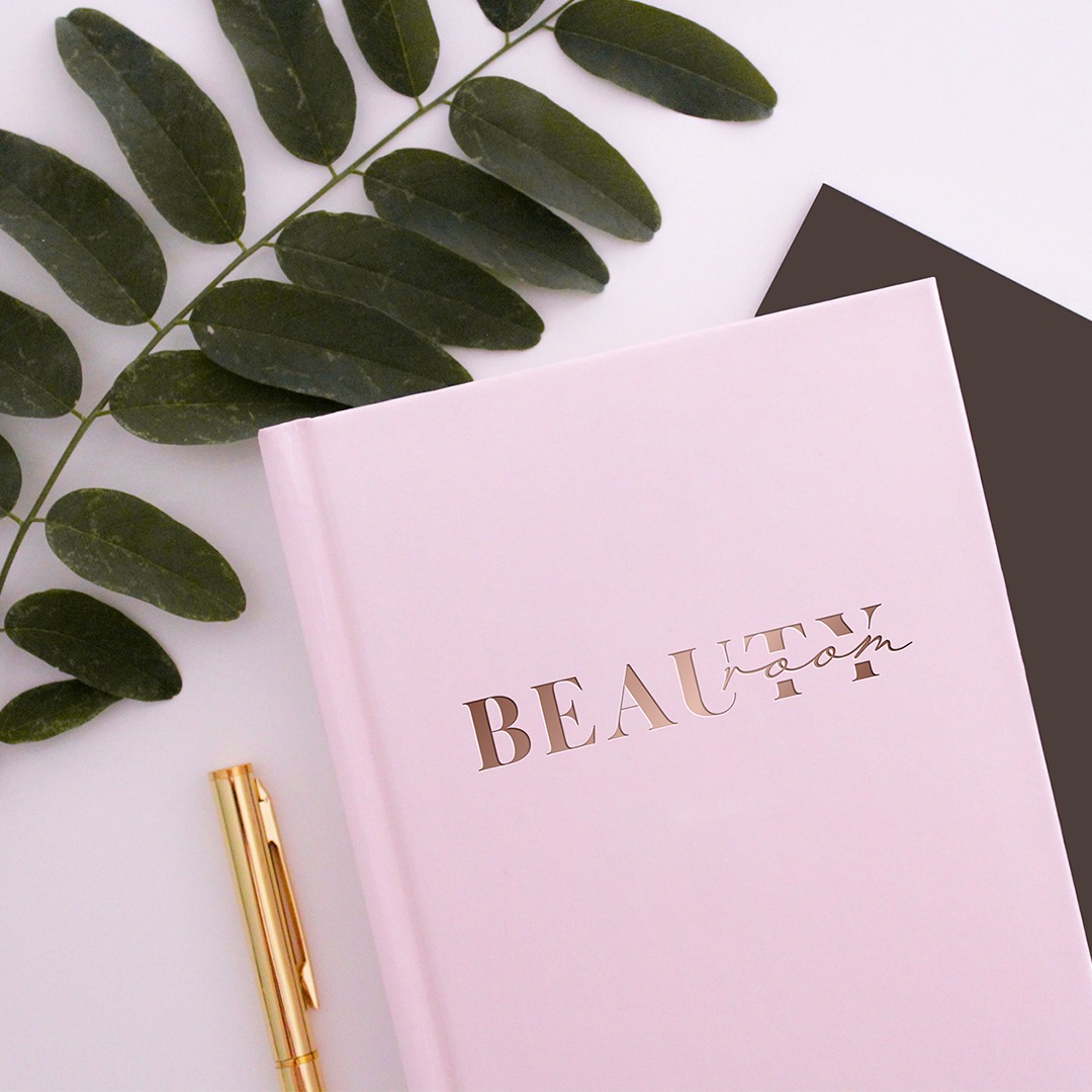 Różowy notatnik z napisem 'BEAUTY room' w kolorze różowego złota, złoty długopis, gałązka zielonych liści i ciemnobrązowa karta na białym tle.