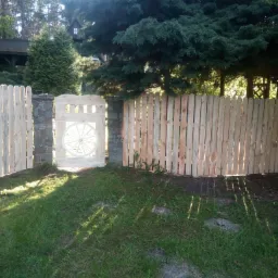 Nowy, jasny płot drewniany z furtką ozdobioną kołem od wozu, wsparty na kamiennych słupkach, otoczony zielonym trawnikiem i drzewami.