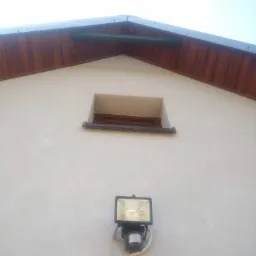 Ściana budynku z oknem i zamontowanym reflektorem z czujnikiem ruchu, brązowe drewniane wykończenie pod dachem, jasny tynk.