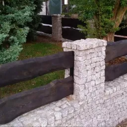 Ogrodzenie z ciemnego drewna i jasnoszarego kamienia, z widocznymi słupkami i poziomymi belkami, otoczone zielenią.