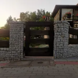 Wejście na posesję z drewnianą bramą i kamiennymi słupkami z kostki, widok z poziomu ulicy.