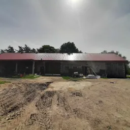 Budynek gospodarczy z nowym, czerwonym dachem z blachy trapezowej, widoczne ślady opon na ziemi, drabina oparta o ścianę.