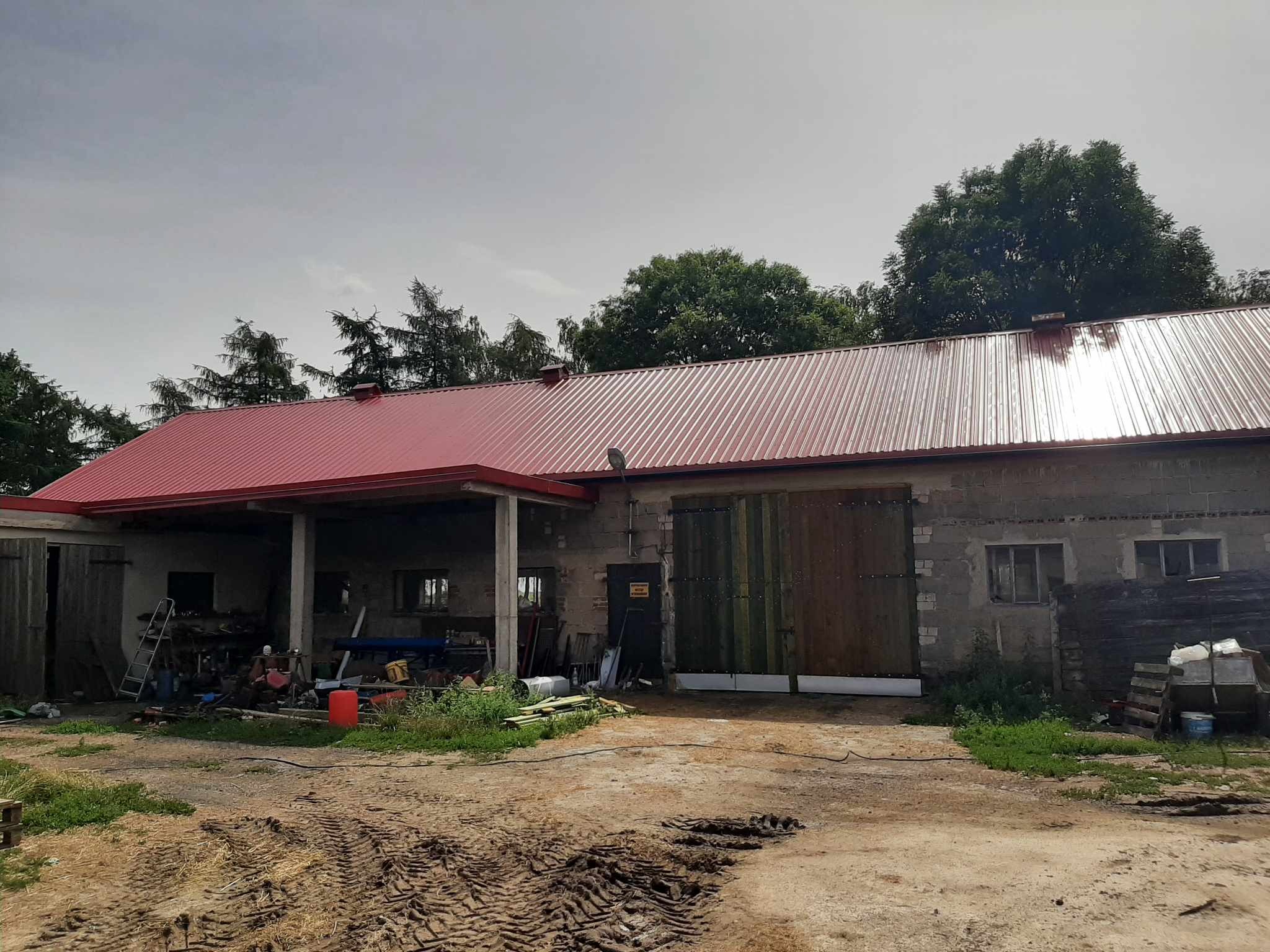 Budynek gospodarczy z czerwonym, metalowym dachem, widoczny z zewnątrz na tle pochmurnego nieba i drzew. Ziemia przed budynkiem poorana śladami opon.