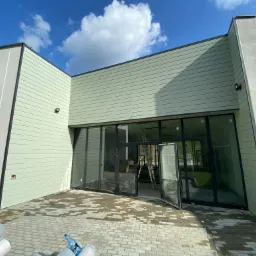 Nowoczesny budynek z elewacją w odcieniach szarości i zieleni, duże szklane okna i drzwi, na zewnątrz kostka brukowa, w tle błękitne niebo z chmurami, widoczna drabina wewnątrz budynku.