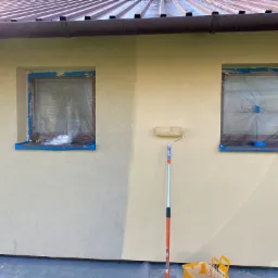 Elewacja budynku w trakcie malowania, okna zabezpieczone folią i taśmą, widoczny wałek malarski i wiadro z farbą, fragment ściany już pomalowany na jaśniejszy odcień.