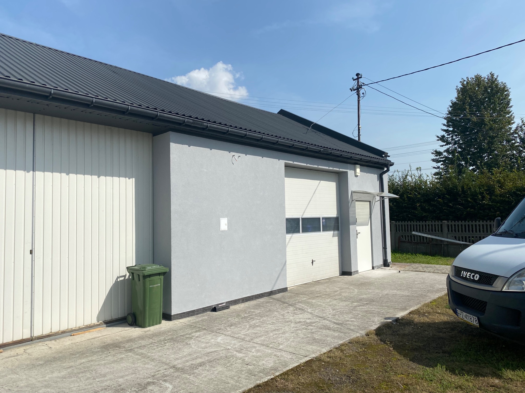 Szary budynek z czarnym dachem i białą bramą garażową, z zielonym koszem na śmieci i zaparkowanym białym samochodem dostawczym na betonowym podjeździe w słoneczny dzień.