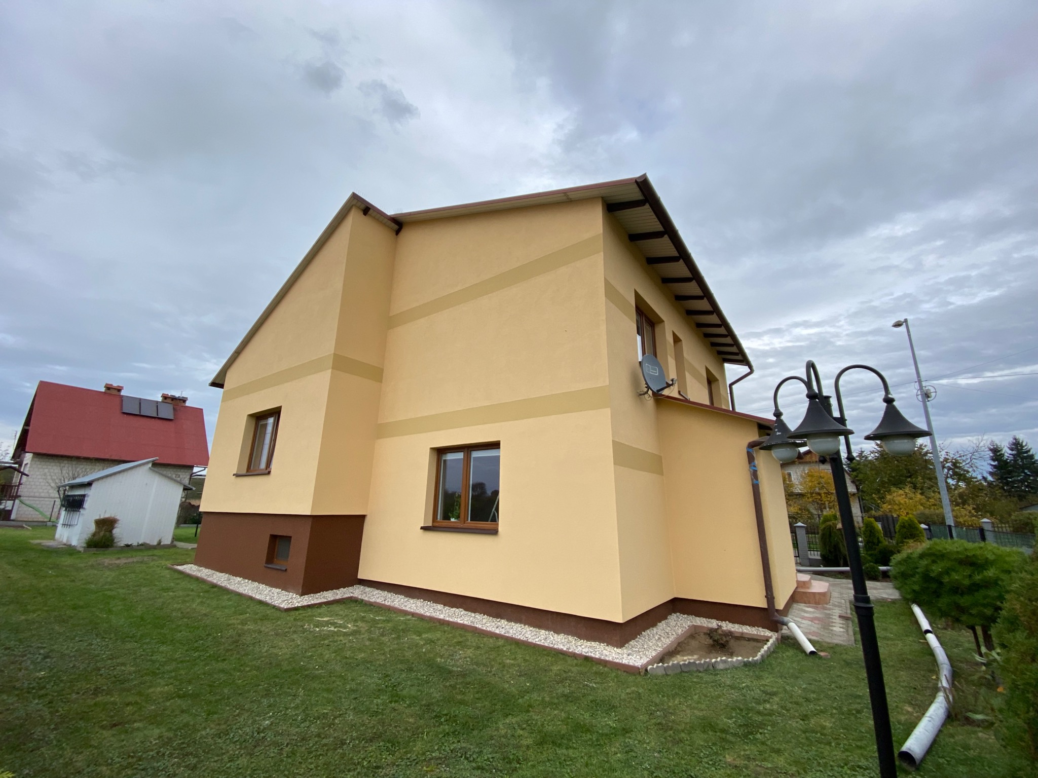 Dwukondygnacyjny dom z beżową elewacją i brązowym cokołem, otoczony zielonym trawnikiem i szarym żwirem, sfotografowany w pochmurny dzień z niskiej perspektywy.