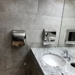 Publiczna toaleta z umywalką z szarego granitu, chromowaną baterią, podajnikiem na ręczniki papierowe i suszarką do rąk ze stali nierdzewnej, wykończona płytkami w odcieniach szarości.