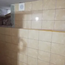 Beżowe płytki ceramiczne na ścianie łazienki, z widoczną kratką wentylacyjną i stertą płytek w tle.