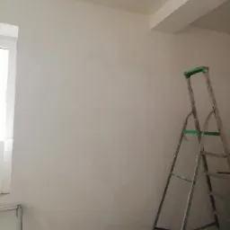 Świeżo wykończona, gładka ściana w jasnym odcieniu z widoczną drabiną i fragmentem okna, ukazująca etap prac wykończeniowych.