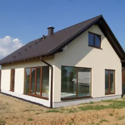 Nowoczesny dom jednorodzinny z brązowym dachem i oknami, kremowa elewacja, widok z zewnątrz na tle błękitnego nieba.