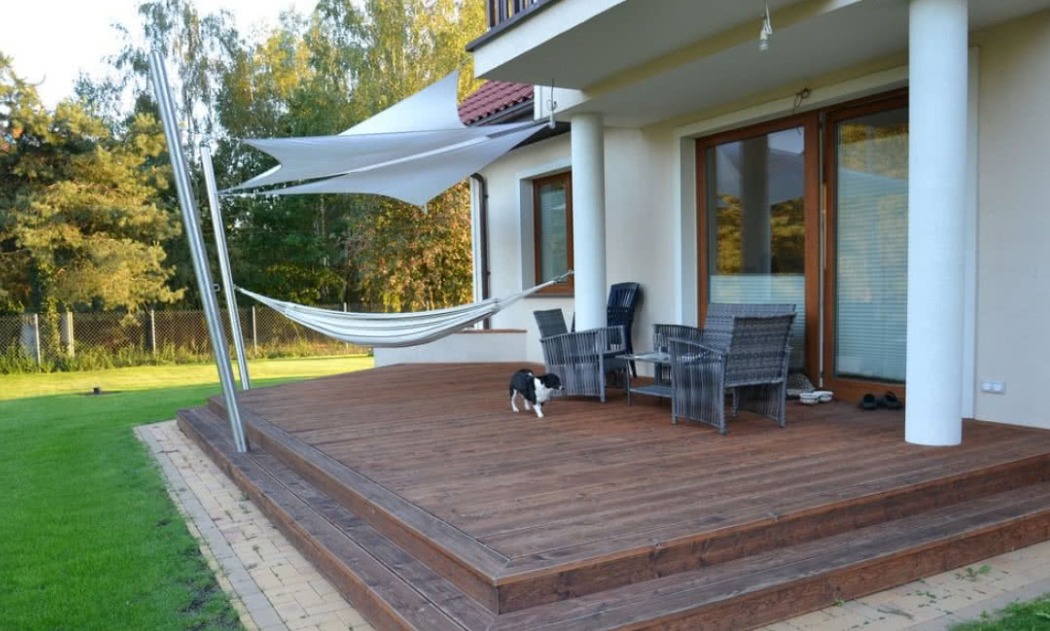 Zadaszony taras z drewnianą podłogą, hamakiem i zacienieniem żaglowym, na którym stoi czarno-biały pies.