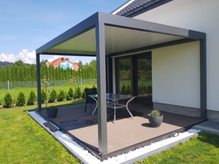 Nowoczesna pergola z szarymi słupami i białym zadaszeniem, z tarasem z desek kompozytowych, otoczona zielonym trawnikiem i białymi kamieniami, z widokiem na ogród.
