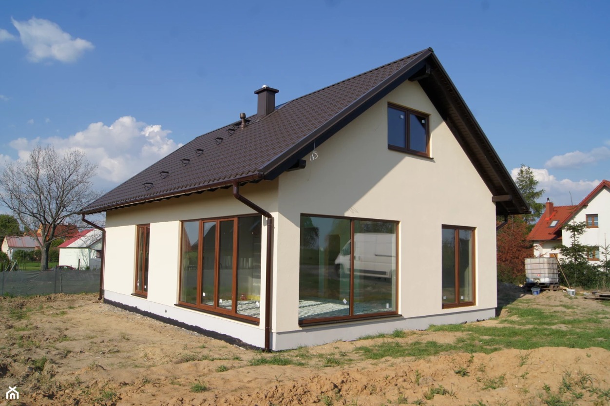 Nowoczesny dom jednorodzinny z brązowym dachem i oknami, kremowa elewacja, widok z zewnątrz na tle błękitnego nieba.