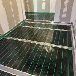 Rozłożona czarna mata grzewcza z zielonymi liniami na podłodze w surowym pomieszczeniu z częściowo wykończonymi ścianami.