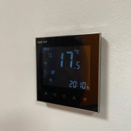 HEAT AT HOME SP. Z O.O. - Czarny, dotykowy termostat marki Heat-On zamontowany na białej, teksturowanej ścianie, wyświetlający temperaturę 17.5 stopni Celsjusza i godzinę 20:10, z ikonami Wi-Fi i ogrzewania.