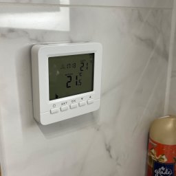 HEAT AT HOME SP. Z O.O. - Biały termostat na marmurowej ścianie, wyświetlający temperaturę pokojową 21.5 stopni Celsjusza i ustawioną na 21 stopni Celsjusza, obok odświeżacz powietrza Glade o zapachu jabłkowego cydru.