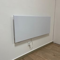 HEAT AT HOME SP. Z O.O. - Płaski, biały panel grzewczy zawieszony na białej ścianie, podłączony do gniazdka elektrycznego, widoczna drewnopodobna podłoga i fragment drzwi.