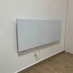 Płaski, biały panel grzewczy zawieszony na białej ścianie, podłączony do gniazdka elektrycznego, widoczna drewnopodobna podłoga i fragment drzwi.