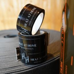 HEAT AT HOME SP. Z O.O. - Dwie rolki czarnej taśmy z logo firmy grzewczej HEAT AT HOME ułożone jedna na drugiej, obok zwiniętej szarej folii grzewczej.
