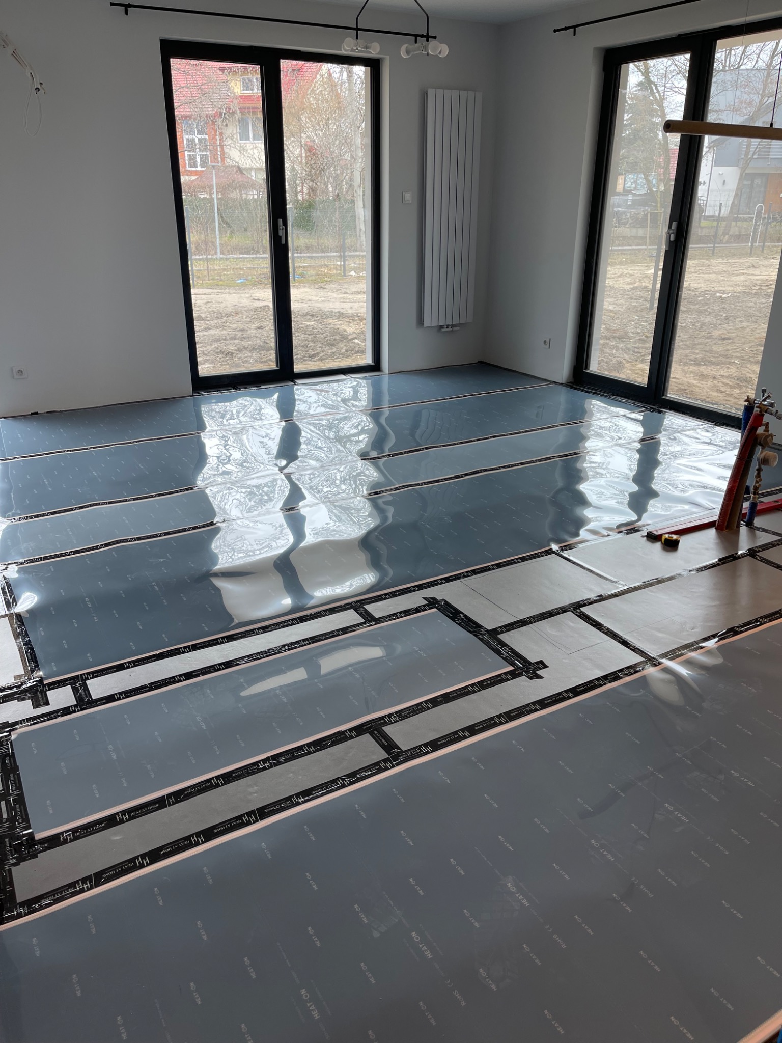 Rozłożone folie grzewcze z widocznym nadrukiem na podłodze w jasnym pomieszczeniu z dużymi oknami, przygotowane do montażu systemu ogrzewania podłogowego.