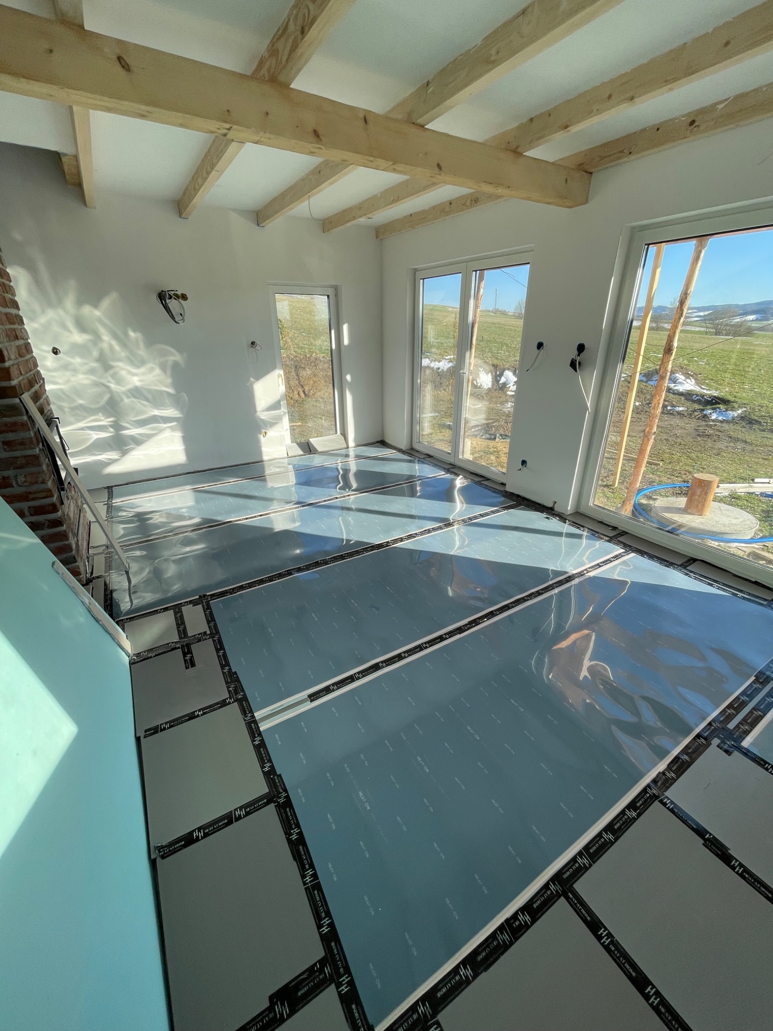 Przygotowanie podłoża pod system grzewczy w jasnym pomieszczeniu z drewnianymi belkami stropowymi i widokiem na zaśnieżony krajobraz za oknem.