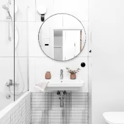 Minimalistyczna, jasna łazienka z okrągłym lustrem, białą umywalką, mozaiką i terazzo na podłodze; akcent kolorystyczny w postaci dozownika na mydło i pojemnika na szczotkę WC w odcieniach terakoty.