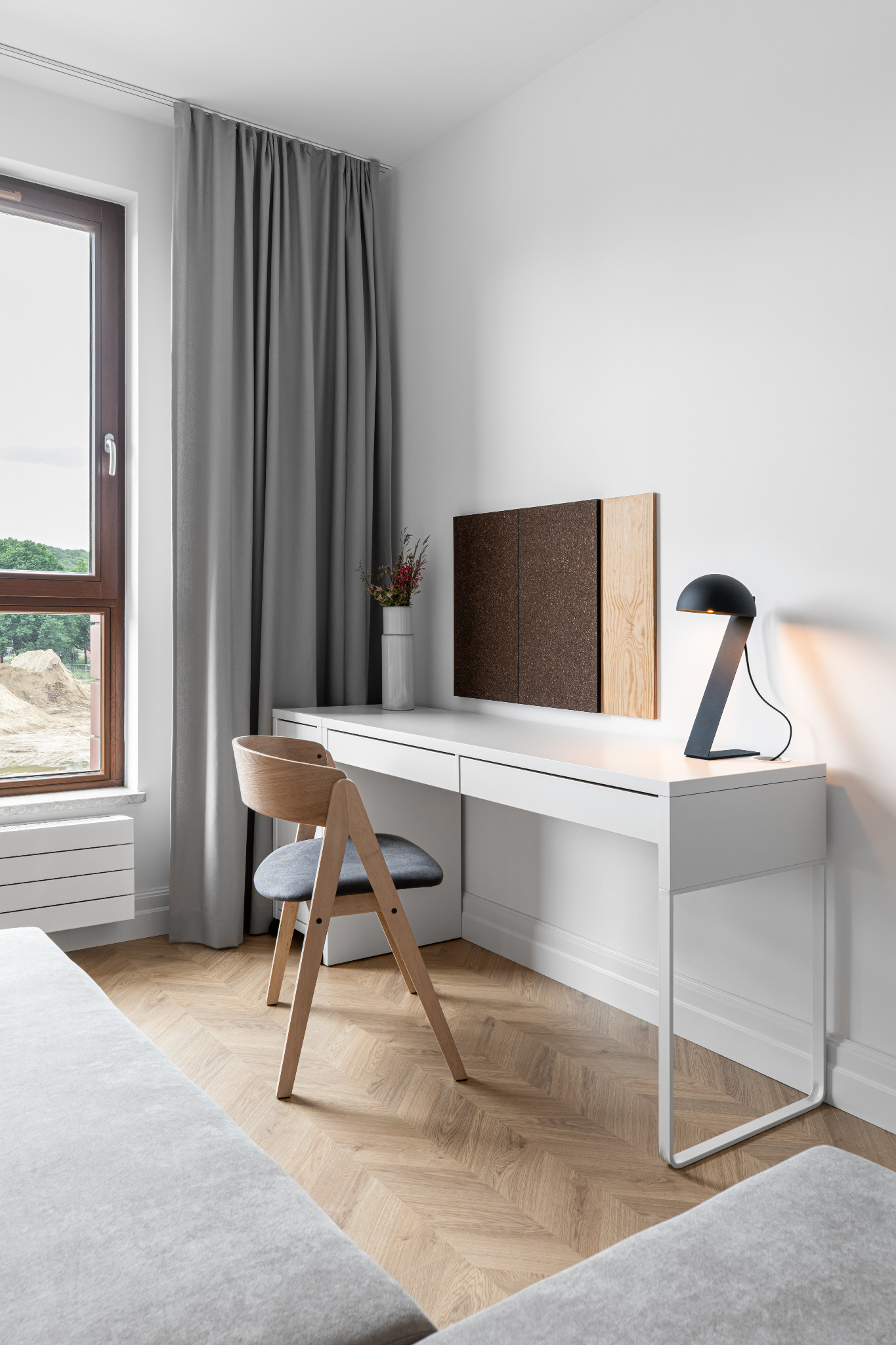 Nowoczesny kącik do pracy z białym biurkiem, drewnianym krzesłem z szarym siedziskiem, korkową tablicą, minimalistyczną lampką i wazonem z kwiatami, na tle parkietu w jodełkę i okna z widokiem...