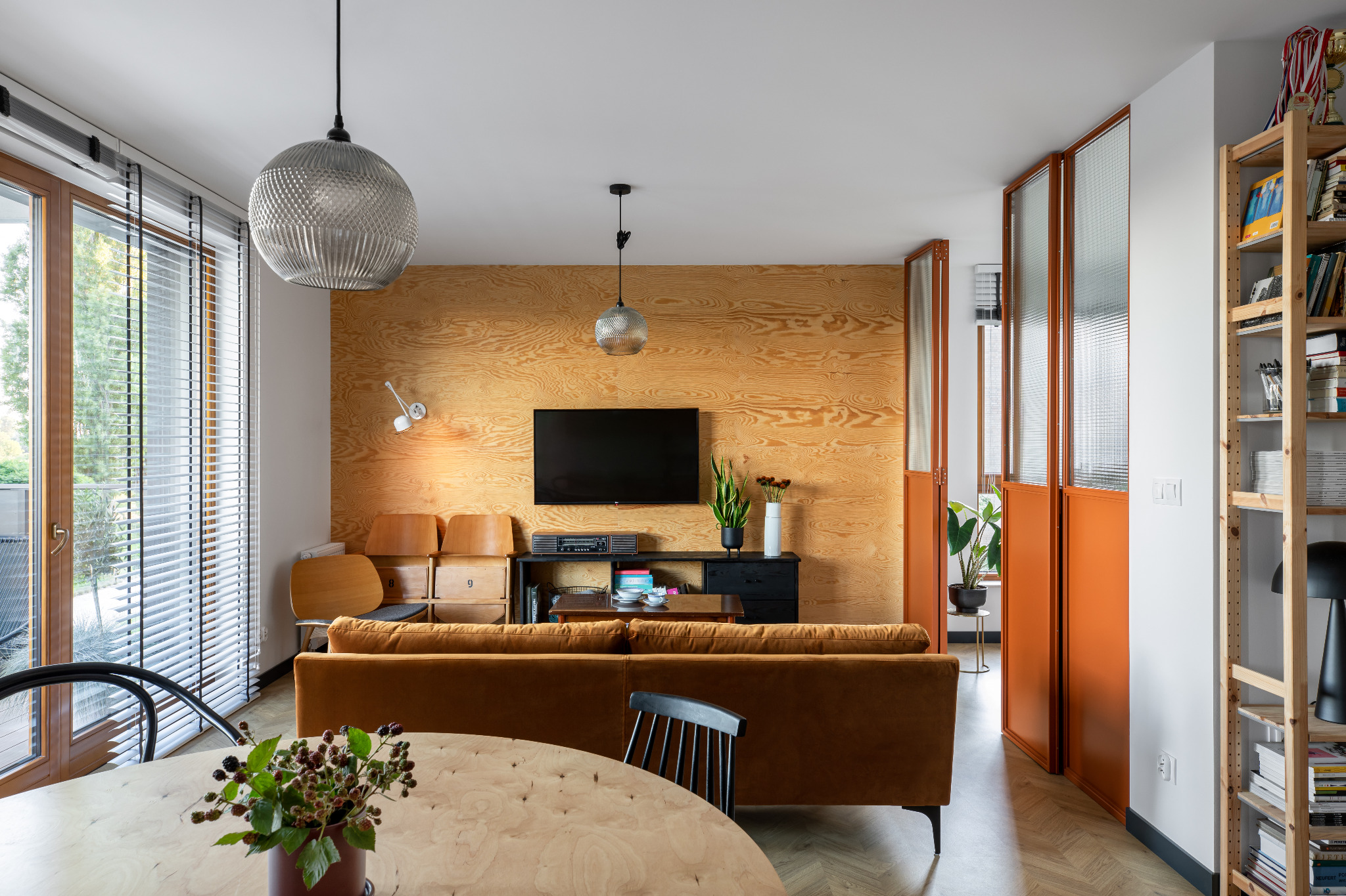 Jasne wnętrze salonu z drewnianą ścianą, brązową sofą, okrągłym stołem i pomarańczowymi, przeszklonymi drzwiami przesuwnymi. Widoczna lampa wisząca w kształcie kuli, drewniany regał z książkami...