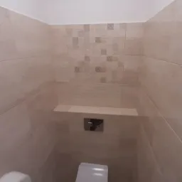 Nowoczesna toaleta z beżowymi płytkami, zabudowaną spłuczką i półką, oraz dyskretnym oświetleniem górnym.