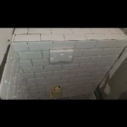 Ściany w trakcie układania białych, prostokątnych płytek z krzyżykami dystansowymi; widoczne rury instalacji wodnej i fragment styropianu w ścianie.