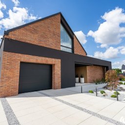 BRAMEO MIESZKO STOŁOSKI - Nowoczesny dom z cegły klinkierowej i antracytową bramą garażową. Minimalistyczny ogród z białymi płytami i szarym żwirem. Błękitne niebo z obłokami w tle.