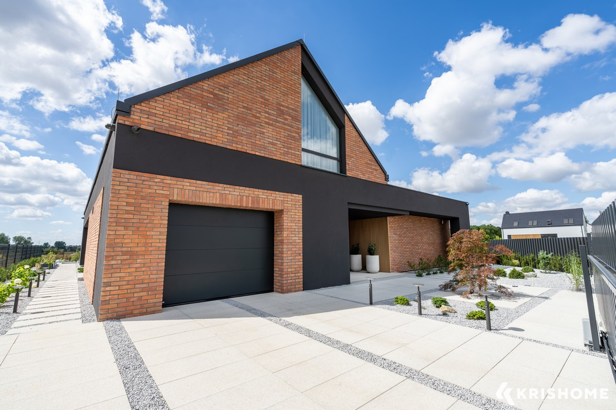 Nowoczesny dom z cegły klinkierowej i antracytową bramą garażową. Minimalistyczny ogród z białymi płytami i szarym żwirem. Błękitne niebo z obłokami w tle.
