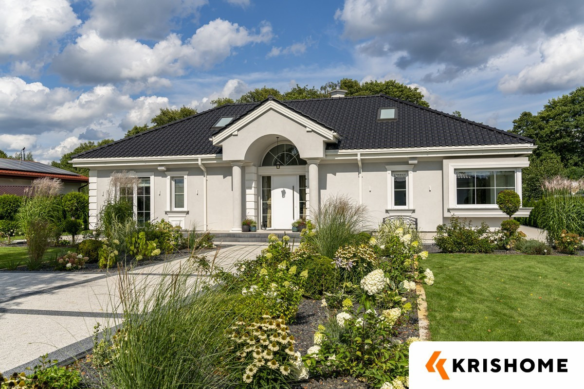 Elegancki, parterowy dom z białymi oknami i drzwiami wejściowymi, otoczony bujnym ogrodem. Widoczne kolumny przy wejściu i czarny dach z lukarnami.