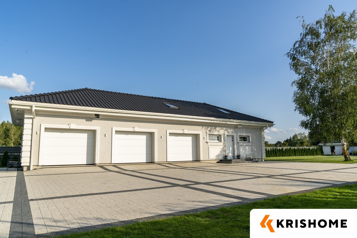 Nowoczesny budynek garażowy z trzema białymi bramami, ciemnym dachem i jasną elewacją, otoczony kostką brukową i zielenią w słoneczny dzień.