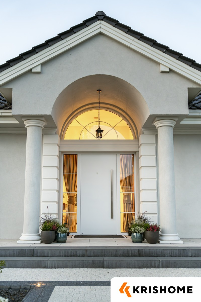 Eleganckie wejście do domu z białymi drzwiami, kolumnami i łukowym oknem nad drzwiami. Widoczne rośliny w donicach i oświetlenie zewnętrzne.