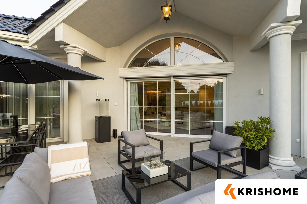 Elegancki taras z meblami wypoczynkowymi i dużymi przesuwnymi oknami z roletą zwiniętą do góry. Widoczne kolumny i oświetlenie zewnętrzne.