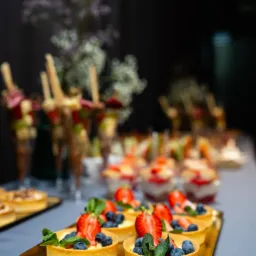 Ekskluzywny catering: mini tartaletki z kremem, borówkami i truskawkami na złotej tacy, w tle desery w szklankach i kwiaty. Elegancka prezentacja oferty 'Luxury'.