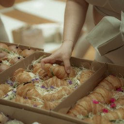 Top Party Box Sp. z o.o. - Kobieta wkłada rogalika do kartonowego pudełka z cateringiem: kanapki, rogaliki z dekoracją z kwiatów i ziół. Pudełka z jasnego kartonu.