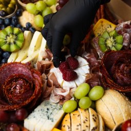 Top Party Box Sp. z o.o. - Elegancki box cateringowy: ręka w czarnej rękawiczce układa maliny na serze brie. W tle sery, wędliny, owoce, pieczywo i pikle, tworzące apetyczną kompozycję.