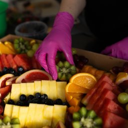 Top Party Box Sp. z o.o. - Ręka w różowej rękawiczce układa plaster kiwi w boxie z owocami: arbuz, ananas, pomarańcze, grejpfruty, winogrona i borówki. Klasyczny catering w boxach.