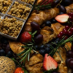 Top Party Box Sp. z o.o. - Bogata kompozycja cateringowa: rogaliki, owoce (winogrona, truskawki, jagody), granola w pojemnikach i gałązki rozmarynu tworzą apetyczną całość.