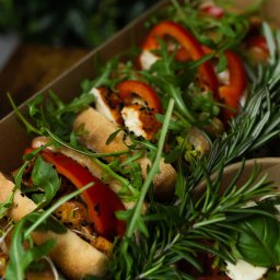 Top Party Box Sp. z o.o. - Kreatywnie zapakowany catering w boxach: kanapki z papryką, rukolą i rozmarynem, widoczne w brązowym pudełku na tle zieleni. Estetyczna kompozycja.