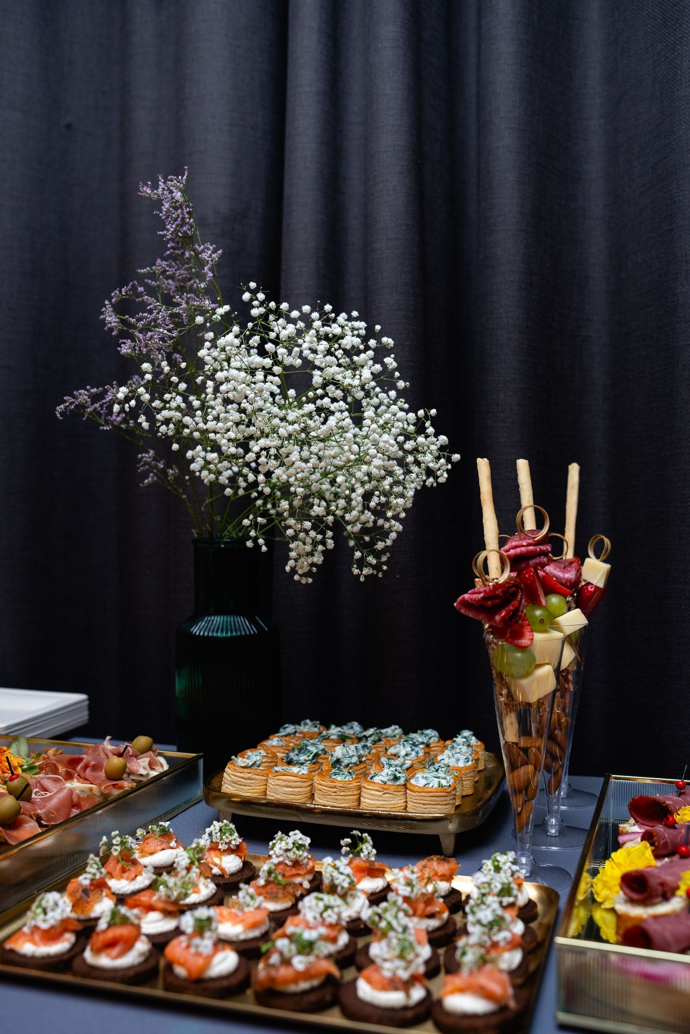 Elegancki stół cateringowy z różnorodnymi przekąskami, kwiatami w wazonie i minimalistycznym tłem. Luksusowa oferta z dbałością o detale.