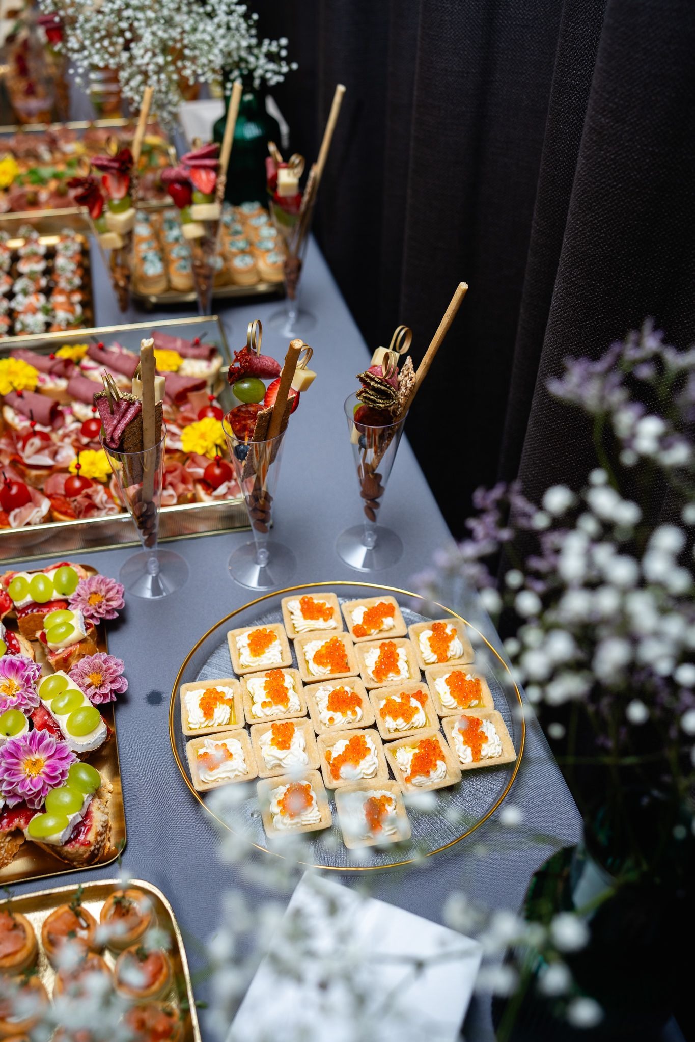 Luksusowy stół cateringowy: kanapeczki z kawiorem na szklanym talerzu, przekąski w kieliszkach, tace z wędlinami i owocami, kwiatowe dekoracje. Elegancka aranżacja.