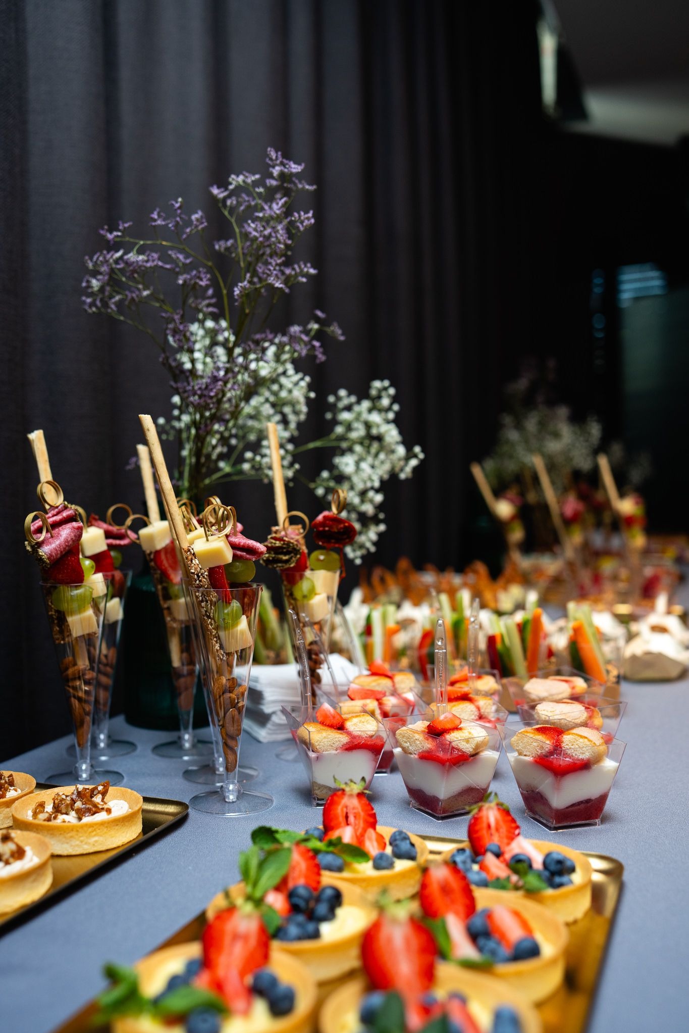 Elegancki stół cateringowy z różnorodnymi przekąskami: mini tartaletki z owocami, deserki w kubeczkach, koreczki owocowo-mięsne i warzywne, dekoracje kwiatowe. 'Oferta Luxury'.